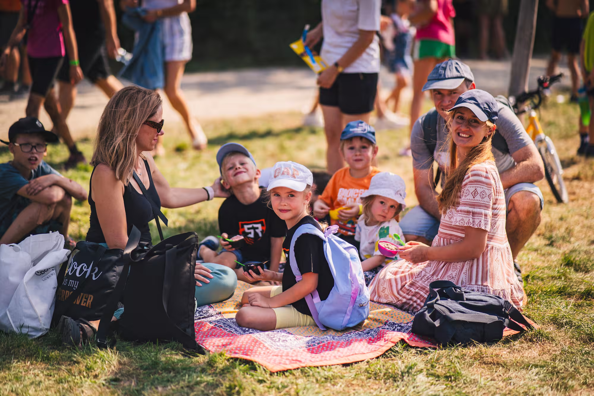 Alternative Text (Tekst alternatywny):Grupa rodziców i dzieci odpoczywa na kocu w strefie piknikowej podczas sportowego wydarzenia Survival Race Kids.